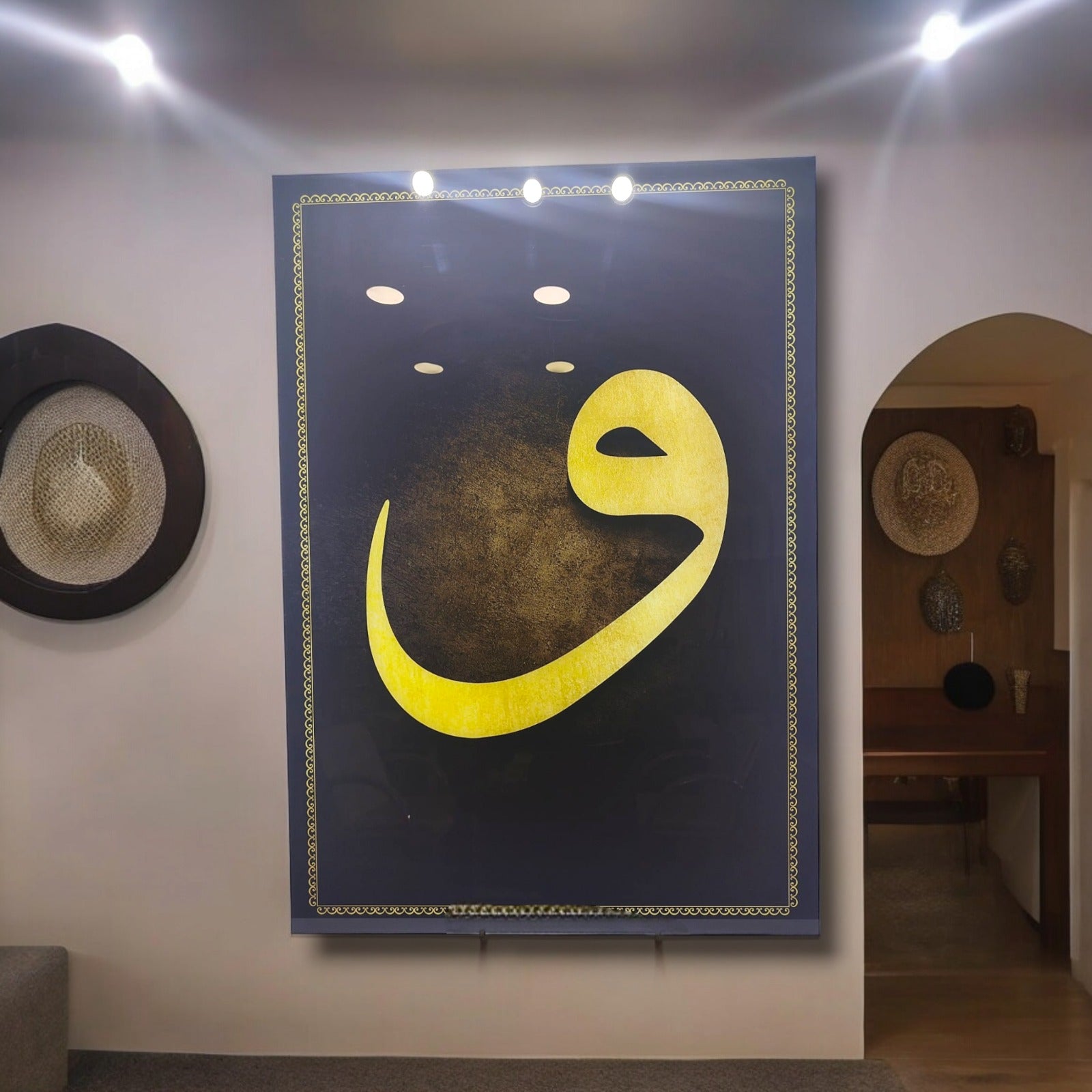 Decorative wall art featuring a bold yellow Arabic letter on a dark background, illuminated by recessed lights.